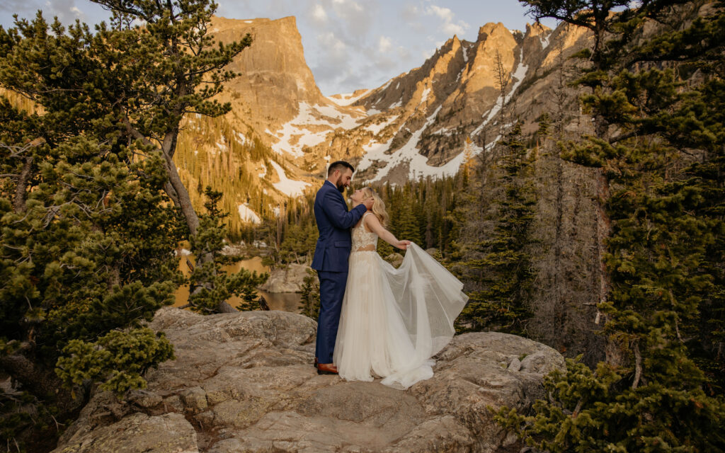 Couple in wedding attire standing above alpine lake at sunrise.
