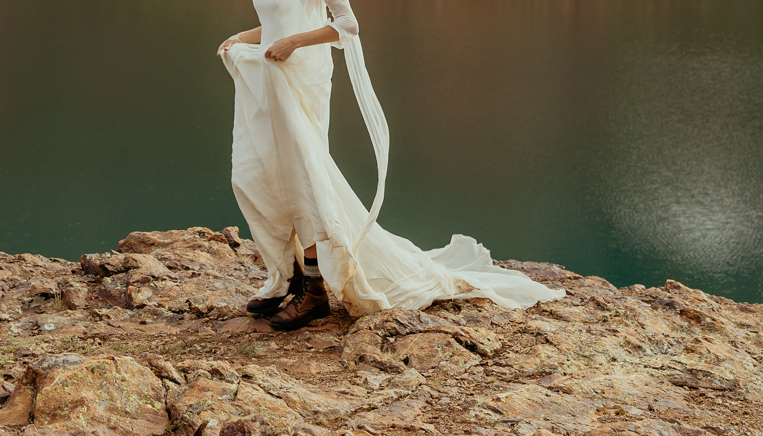 Cropped image of bride in flowy dress with hiking boots by alpine lake.