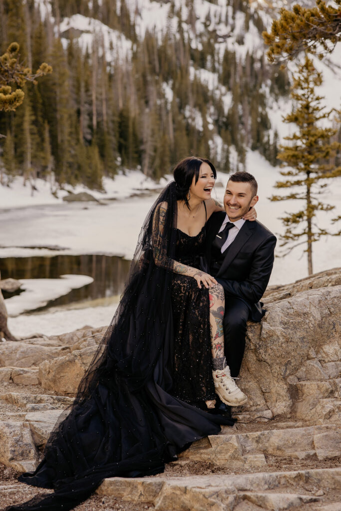 Non-traditional couple sitting on rock in black elopement wedding dress and black suit.