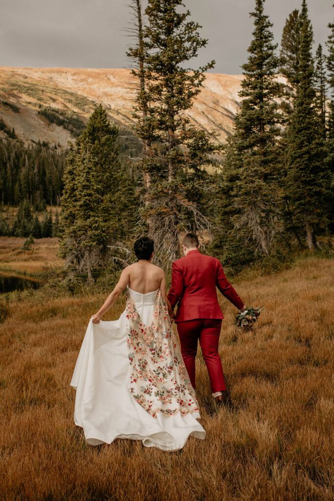 Non-traditional couple walking in field wearing white wedding dress with embroidered cape and red suit.