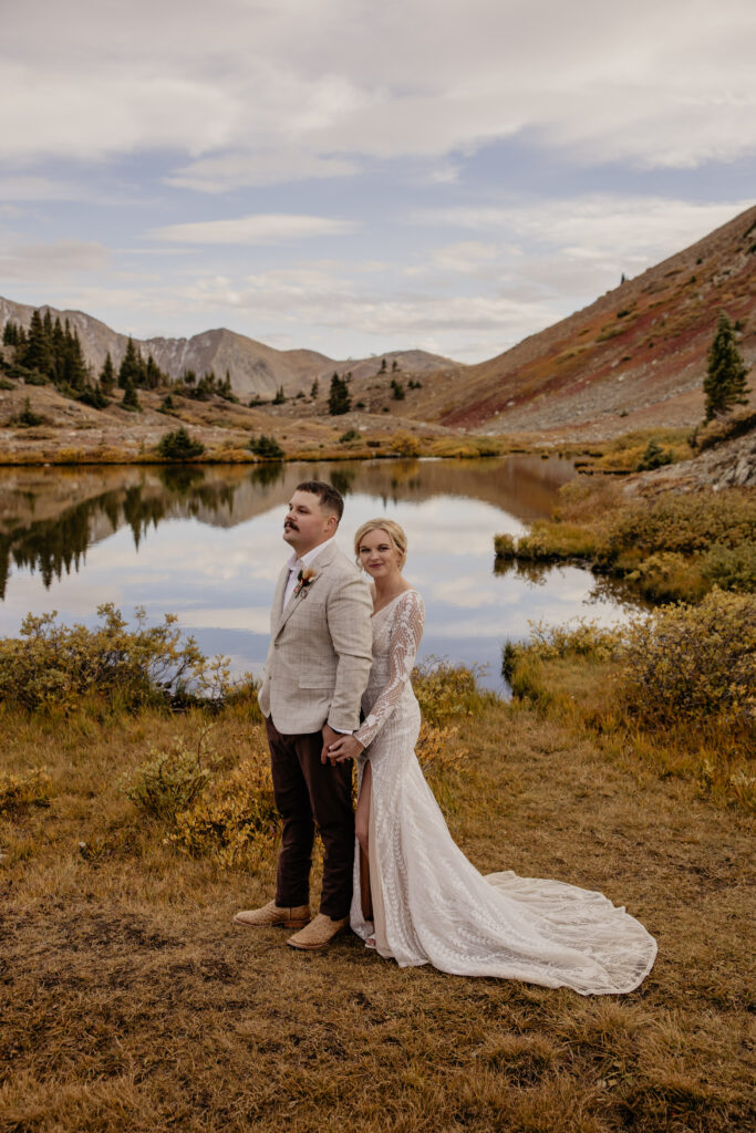 Wedding couple poses near alpine lake with mountains in the background.