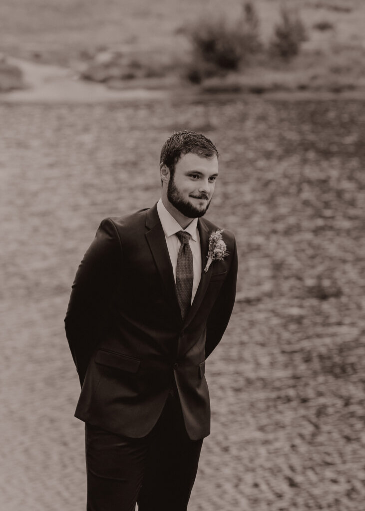 Black and white photo of groom softly smiling at bride as she walks down path.