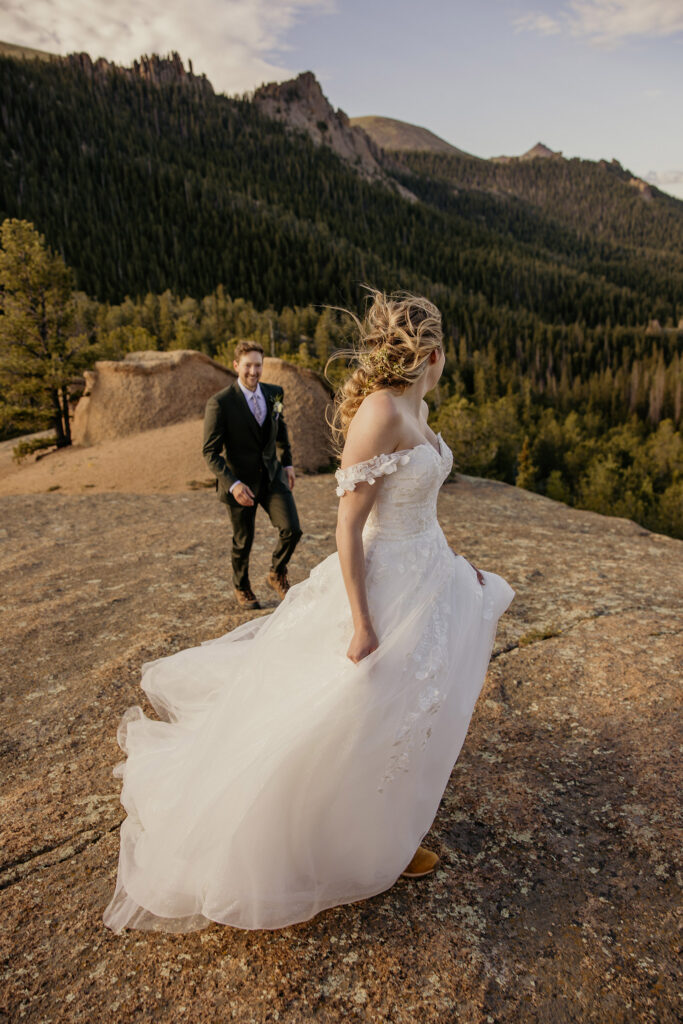 Bride running while looking behind her at groom with mountains in background.