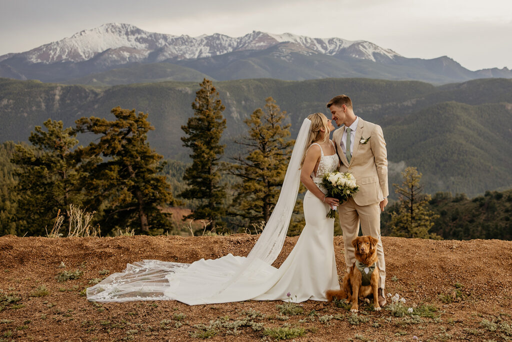Wedding couple standing side by side looking at each other with their dog at their feet and Pikes Peak in the background.