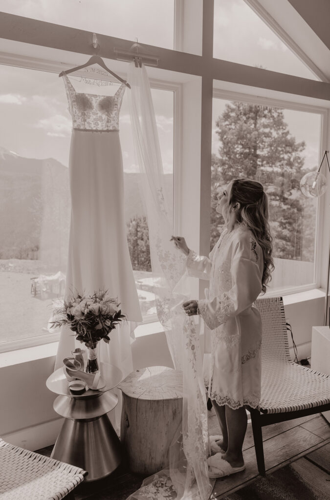 Bride admires her dress hanging in the window before her wedding day.
